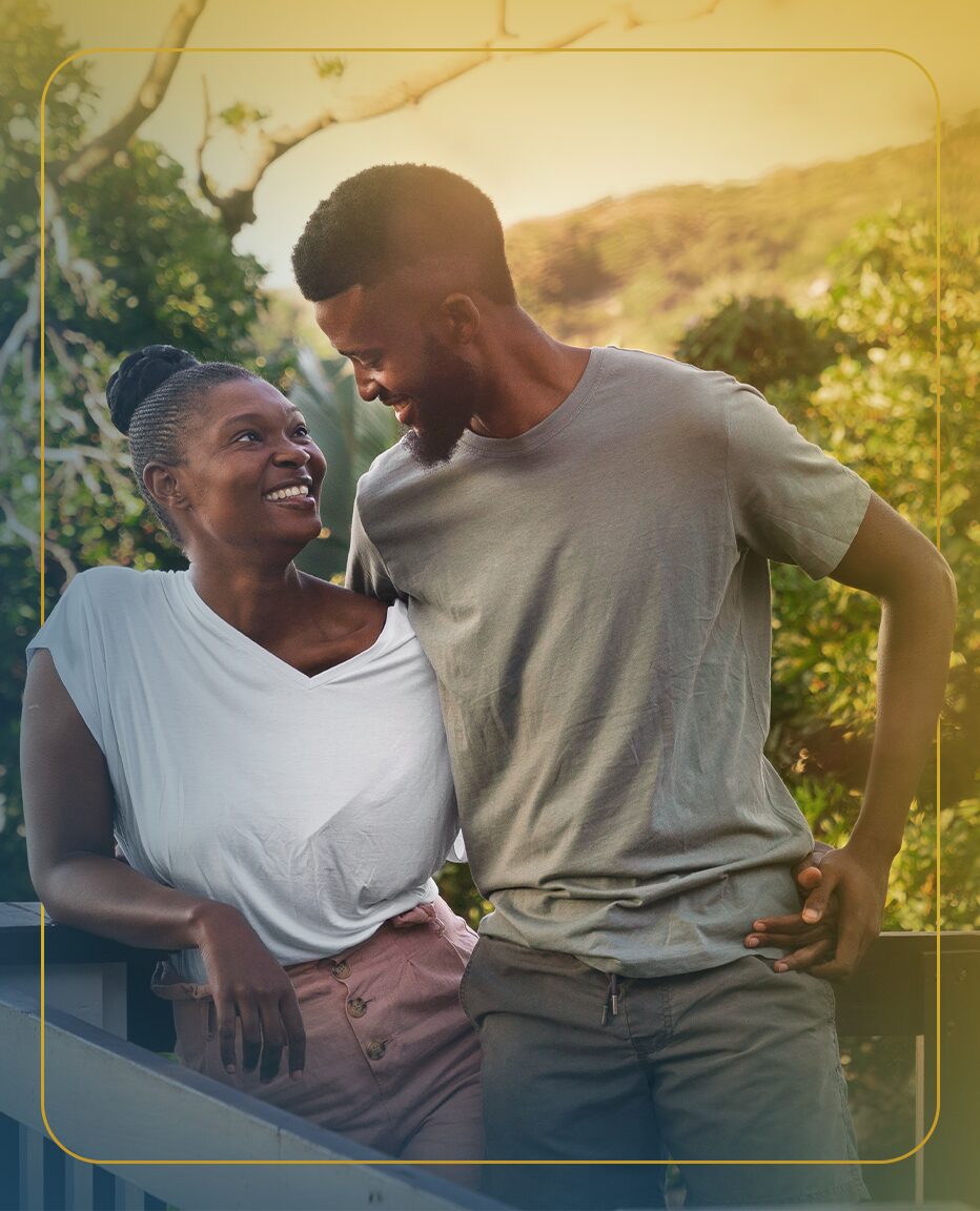 A couple stands close together outside their lake house, as the woman rests her head on her husband's shoulder.